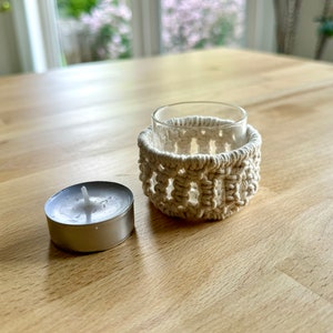 Peut inclure: Un bougeoir en macramé blanc avec un verre à bougie à l'intérieur. Une bougie chauffe-plat argentée repose sur une table en bois.