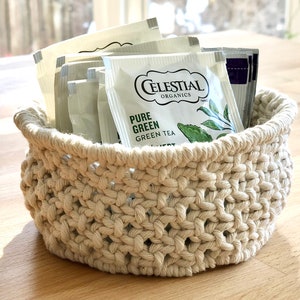 Peut inclure: Un panier en crochet beige rempli de sachets de thé. Les sachets de thé sont étiquetés "Celestial Organics Pure Green Green Tea".