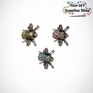 May include: Three bee-shaped brooches with rhinestone accents. The brooches are silver-toned and feature different colored rhinestones: yellow, pink, and blue.