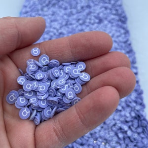 May include: A handful of small, purple, round, flat, plastic, resin, or clay charms with a white Taurus zodiac symbol printed on each charm.