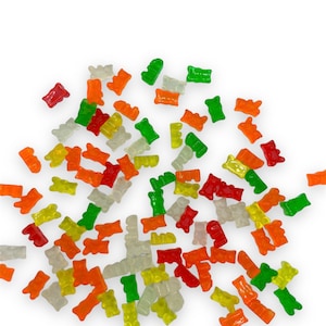Op de afbeelding: Een grote verzameling gummy bears in verschillende kleuren, waaronder groen, oranje, rood en wit. De gummy bears zijn verspreid over een witte achtergrond.