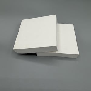 Peut inclure: Deux carreaux de céramique blancs et carrés sont empilés en biais sur un fond gris. Les carreaux ont une finition lisse et mate, probablement à des fins de construction ou de design.