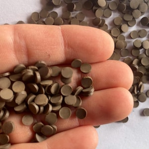 Pode incluir: Uma imagem em close-up mostra um punhado de pequenos objetos marrons, planos e circulares. Alguns são segurados na mão, outros estão espalhados em uma superfície branca. Os objetos são uniformes em tamanho e forma.