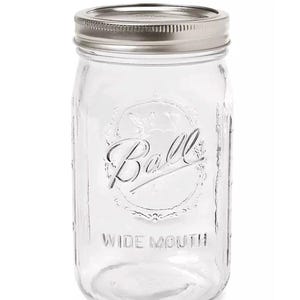 Op de afbeelding: Een heldere glazen Ball wide-mouth pot met een zilveren metalen deksel. De pot heeft in reliëf de tekst "Ball" en "WIDE MOUTH". De pot is ontworpen voor het inmaken en bewaren van voedsel.