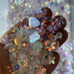 May include: A handful of iridescent sequins and embellishments in various shapes, including seashells, stars, hearts, and flowers. The sequins are in shades of white, pink, and blue, creating a shimmering effect. Ideal for crafting and embellishing projects.