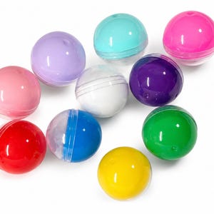 May include: A collection of ten spherical plastic capsules in various colors including pink, lavender, light blue, white, dark purple, red, blue, green, yellow, and hot pink. Each capsule features a clear seam where the two halves join together.
