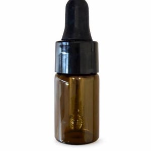 Op de afbeelding: Een klein bruin glazen flesje met een zwarte druppelpipet. De fles is cilindrisch en lijkt leeg te zijn. De druppelpipet heeft een zwarte rubberen bal. De achtergrond is effen wit.