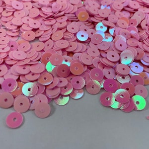 May include: A pile of iridescent pink sequins with a hole in the center. The sequins are scattered on a white surface.