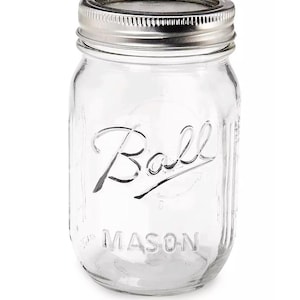 Puede incluir: Un tarro de cristal transparente Ball Mason con tapa de metal plateado. El tarro tiene la palabra "Ball" en cursiva y "MASON" impresa debajo. Diseñado para envasar y conservar alimentos.