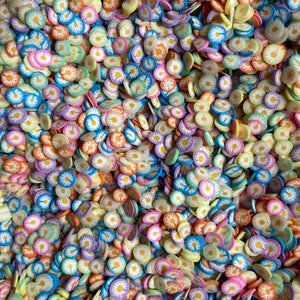 May include: A large quantity of colorful daisy-shaped sprinkles. The sprinkles are in various shades of blue, yellow, pink, orange, and green. Each sprinkle has a white center with a yellow daisy.