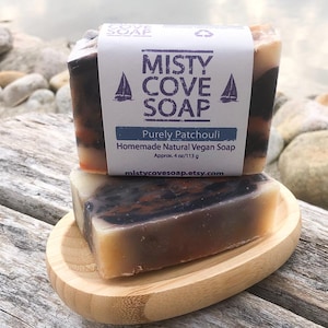 May include: Two bars of handmade natural vegan soap with a black, orange, and white swirl design. The soap is labeled "Misty Cove Soap" and "Purely Patchouli" with a sailboat graphic. The soap is approximately 4 ounces or 113 grams.