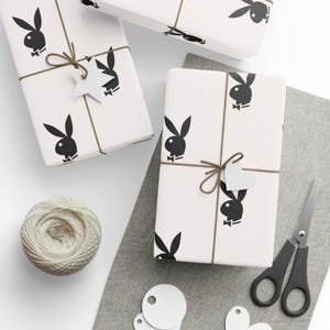 May include: Wrapped gifts with a repeating black bunny logo on white paper, tied with twine and decorated with star and heart-shaped tags. A ball of twine, scissors, and blank tags are also present.