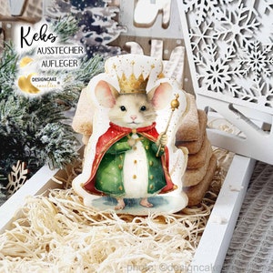 Könnte beinhalten: Ein dekorierter Keks in Form eines Mäusekönigs, der eine Krone und ein Zepter trägt. Die Maus trägt einen roten und grünen Umhang. Der Keks liegt auf einem weißen Tablett mit anderen Keksen und dekorativem Stroh.