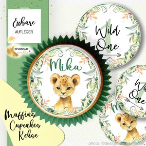 Könnte beinhalten: Ein Set essbarer Cupcake-Topper mit Dschungel-Motiv. Die Topper sind mit einem Löwenjungen, grünen Blättern und dem Text "Wild One" verziert.