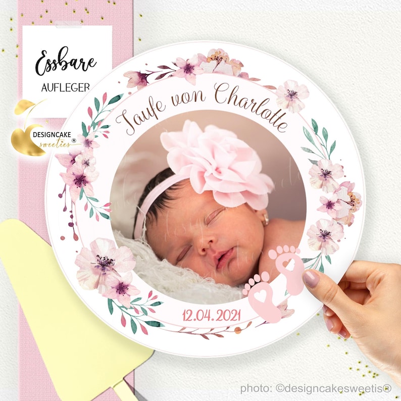 Könnte beinhalten: Ein runder essbarer Kuchentopper in Weiß mit einem rosafarbenen Blumenkranz und einem Fotorahmen. Der Rahmen zeigt ein Foto eines schlafenden Baby-Mädchens mit einem rosafarbenen Blumenhaarband. Der Text "Taufe von Charlotte" und "12.04.2021" ist auf dem Topper gedruckt.