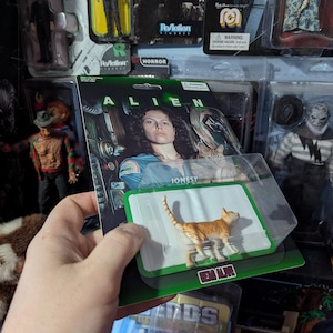 Puede incluir: Figura de acci&oacute;n de un gato atigrado naranja de la pel&iacute;cula Alien, empaquetada en una tarjeta verde y blanca. La tarjeta presenta el t&iacute;tulo de la pel&iacute;cula y el nombre "Jonesy". Otras figuras de acci&oacute;n son visibles en el fondo.