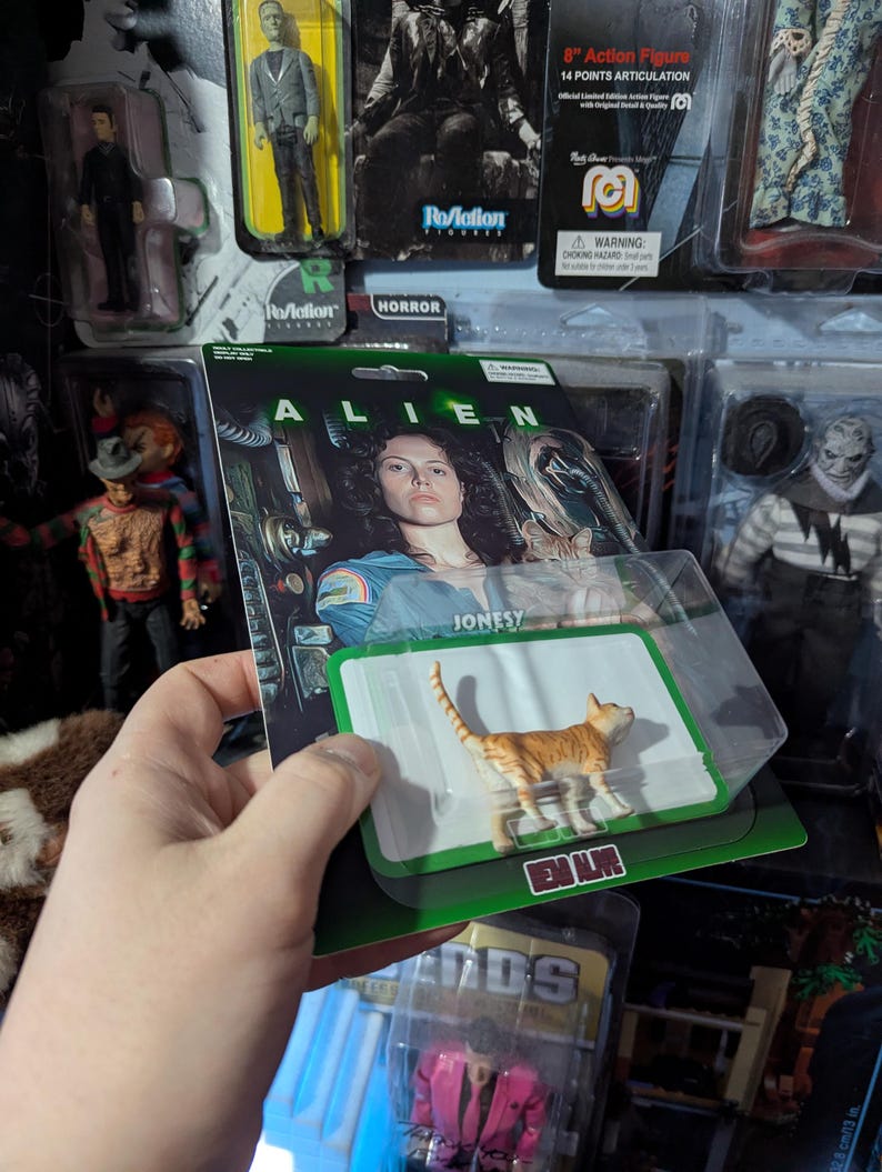 Puede incluir: Figura de acci&oacute;n de un gato atigrado naranja de la pel&iacute;cula Alien, empaquetada en una tarjeta verde y blanca. La tarjeta presenta el t&iacute;tulo de la pel&iacute;cula y el nombre "Jonesy". Otras figuras de acci&oacute;n son visibles en el fondo.