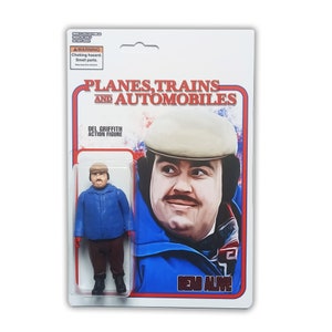 May include: A Del Griffith action figure from Planes, Trains and Automobiles in original blister packaging. The figure wears a blue jacket, brown trousers, and a tan cap. The card features a portrait of the character and the movie title text.