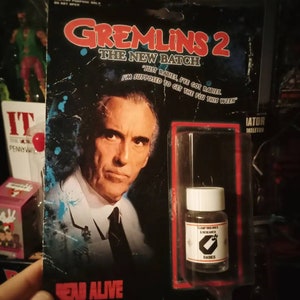 以下が含まれることがあります： 映画「グレムリン2」のコレクター向けアクションフィギュアのパッケージ。男性の肖像画と「狂犬病」と書かれた小さなボトルが描かれています。黒いパッケージには、「グレムリン2：ザ・ニュー・バッチ」と「デッド・アライブ」などの赤と青の文字が書かれています。