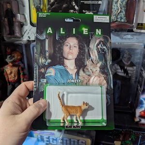 Puede incluir: Figura de acci&oacute;n de un gato de la pel&iacute;cula Alien, con el nombre "Jonesy" en el embalaje. La figura es naranja y marr&oacute;n. El embalaje presenta una foto de la estrella de la pel&iacute;cula y la palabra "ALIEN".