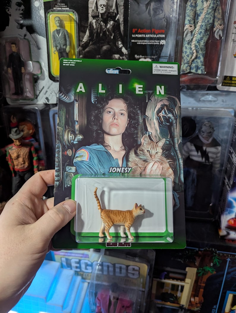 Puede incluir: Figura de acci&oacute;n de un gato de la pel&iacute;cula Alien, con el nombre "Jonesy" en el embalaje. La figura es naranja y marr&oacute;n. El embalaje presenta una foto de la estrella de la pel&iacute;cula y la palabra "ALIEN".