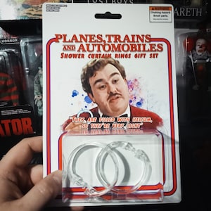 May include: A gift set of shower curtain rings featuring a retro design with the text "PLANES, TRAINS AND AUTOMOBILES". The packaging includes an image of a man and the quote "THEY ARE FILLED WITH HELIUM, SO THEY'RE VERY LIGHT". The rings are clear plastic.