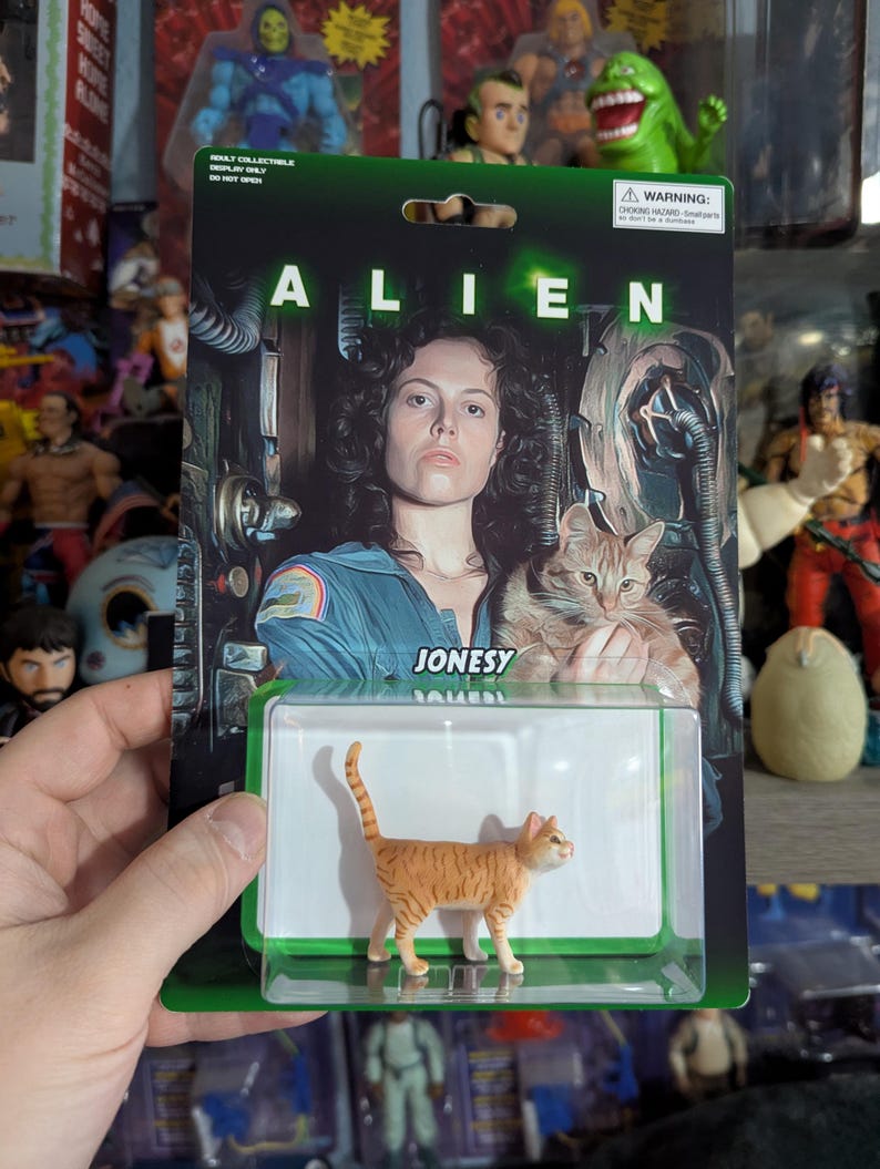 Puede incluir: Figura de acci&oacute;n coleccionable de Jonesy el gato de la pel&iacute;cula Alien. La figura es de color naranja y blanco, de pie en una caja de pl&aacute;stico transparente. El embalaje presenta una foto de la estrella de la pel&iacute;cula y la palabra "ALIEN".