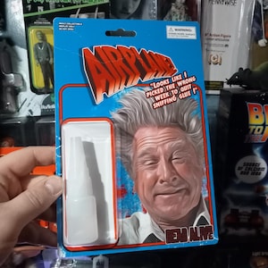 May include: A novelty collectible item with the word "AIRPLANE" in red and white lettering. The packaging features a portrait of a smiling man and the text "LOOKS LIKE I PICKED THE WRONG WEEK TO QUIT SNIFFING GLUE!". A small bottle of glue is visible.