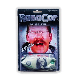 May include: RoboCop "Dollar Play Set" collectible. The packaging features a distressed design with the RoboCop logo and a portrait of a man with glasses. The set includes a crumpled one-dollar bill. Text reads "I'D BUY THAT FOR A DOLLAR!"