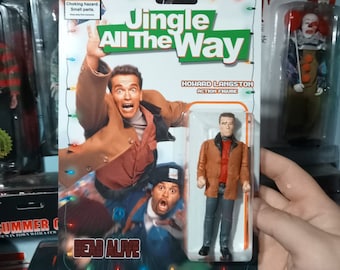 May include: An action figure of Howard Langston from the movie "Jingle All The Way" in a clear plastic package. The packaging features a movie poster with Arnold Schwarzenegger and Sinbad. The text "Jingle All The Way" is displayed prominently.