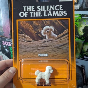 May include: A collectible action figure in orange and black blister packaging titled The Silence of the Lambs. The card features an image of a white dog and the name Precious. A small white dog figurine is enclosed in the clear plastic bubble.