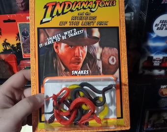 Könnte beinhalten: Ein Indiana Jones Jäger des verlorenen Schatzes Actionfiguren-Set auf einer gelben Blisterkarte mit dem Text "Snakes... why'd it have to be snakes?" und "Snakes". Die durchsichtige Kunststoffblase enthält mehrere kleine, gewundene Kunststoffschlangen in Rot, Gelb und Schwarz.