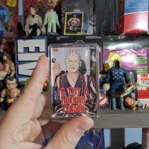 May include: A clear rectangular keychain with a character's face and the text "I'M NOT A MORNING PERSON". The background shows action figures and collectibles. The keychain is held in front of a shelf with various items.