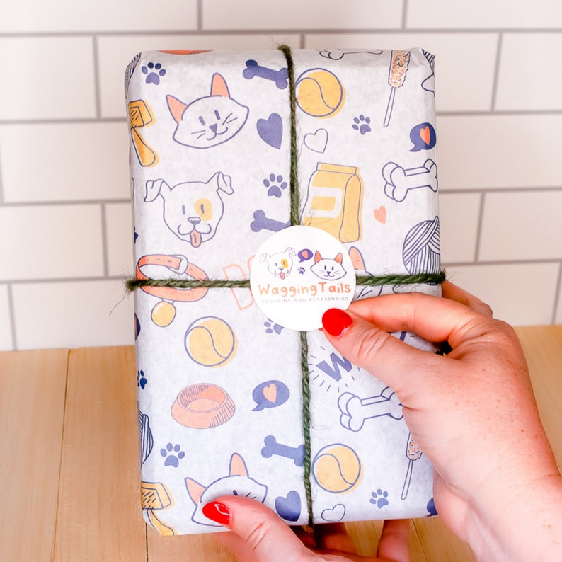 May include: A white gift wrap with a dog and cat pattern. The wrapping paper features paw prints, bones, dog bowls, and toys. A green and brown twine bow is tied around the package. A round sticker with the text "Wagging Tails" is on the front of the package.