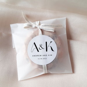 May include: White favor bag with a white circle sticker that says "A&K" in black script font. The sticker also says "Andrew and Kim" and "12.10.2022" in black text.