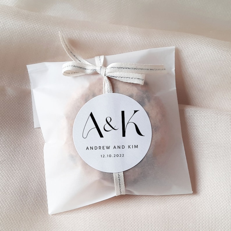 May include: White favor bag with a white circle sticker that says "A&K" in black script font. The sticker also says "Andrew and Kim" and "12.10.2022" in black text.