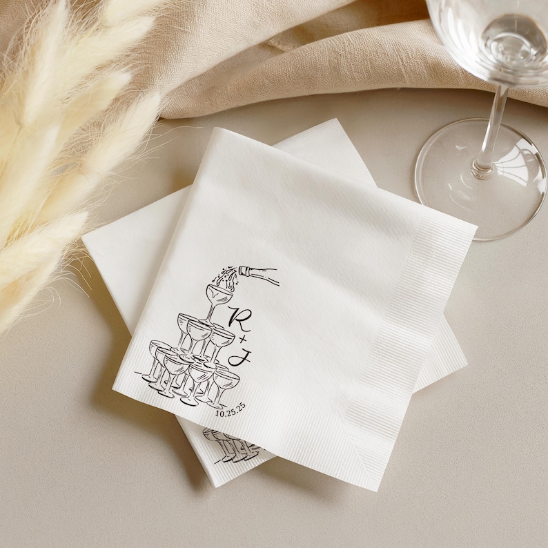 K&ouml;nnte beinhalten: Ein Stapel wei&szlig;er Papierservietten mit einer schwarzen Tuschezeichnung eines Champagnerturms, der aus einer Flasche eingeschenkt wird. Das Design enth&auml;lt die Buchstaben R + J und das Datum 10.25.25 in einer einfachen Serifenschrift.