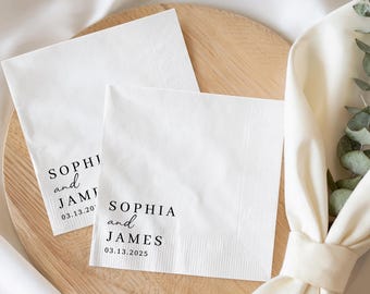 May include: Two white paper napkins with black text that reads "SOPHIA and JAMES" and a date. The napkins are on a wooden tray with a white cloth and greenery in the background. One napkin has the date 03.13.2020 and the other has 03.13.2025.
