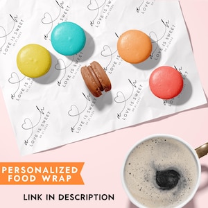K&ouml;nnte beinhalten: F&uuml;nf bunte Macarons in Gelb, T&uuml;rkis, Orange, Braun und Rot auf wei&szlig;em Papier, bedruckt mit dem wiederkehrenden schwarzen Text LOVE IS SWEET und dem Datum 09.21.2025, dazu eine wei&szlig;e Keramiktasse gef&uuml;llt mit schwarzem Kaffee.