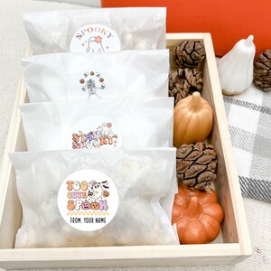 Op de afbeelding: Vier witte papieren zakken met kleurrijke Halloween-stickers. De stickers tonen geesten, pompoenen en skeletten. De zakken bevinden zich in een houten bak met decoratieve pompoenen en dennenappels.