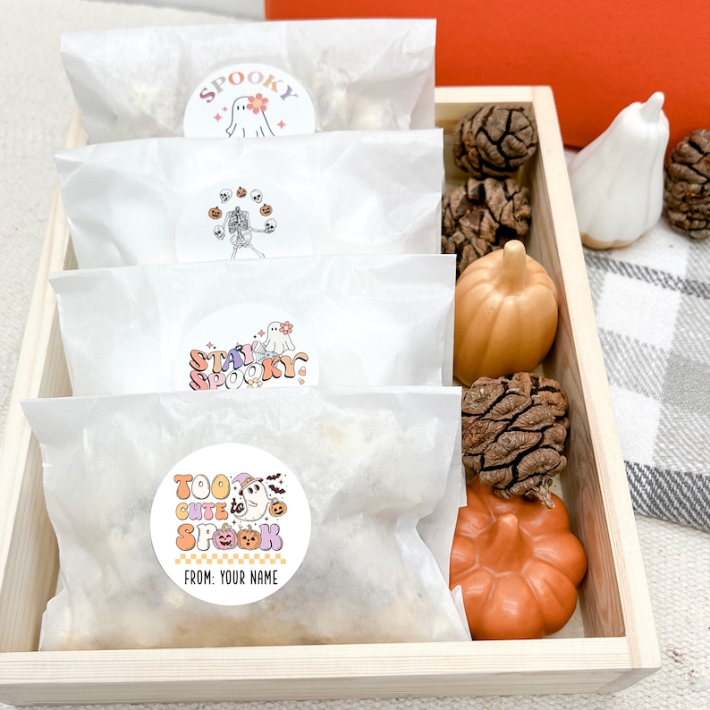 Op de afbeelding: Vier witte papieren zakken met kleurrijke Halloween-stickers. De stickers tonen geesten, pompoenen en skeletten. De zakken bevinden zich in een houten bak met decoratieve pompoenen en dennenappels.
