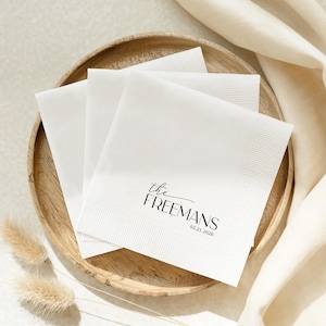 K&ouml;nnte beinhalten: Ein Stapel wei&szlig;er Papierservietten mit schwarzem Aufdruck, der the FREEMANS und das Datum 03.21.2026 zeigt. Die Servietten haben einen strukturierten Rand und liegen auf einem runden Holztablett, daneben ist ein cremefarbener Stoff drapiert.