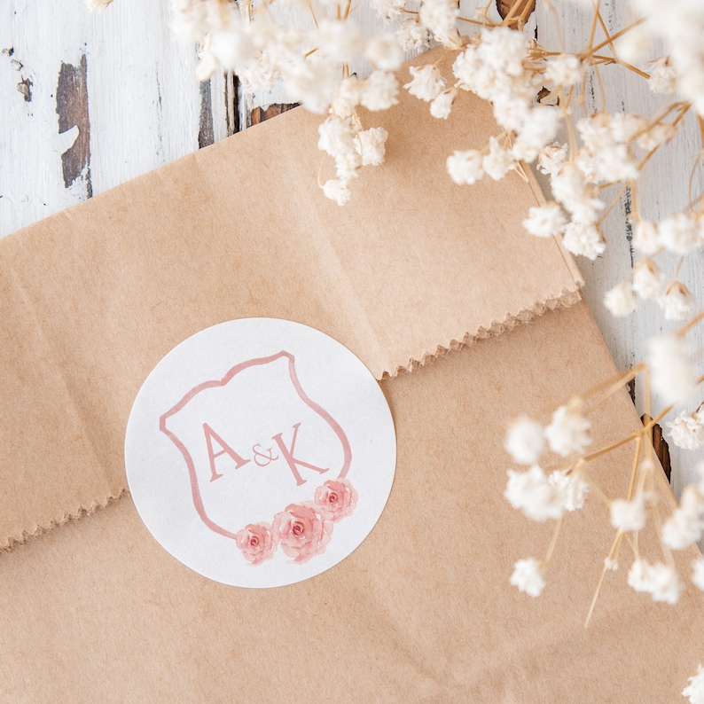 May include: A white round sticker with a pink shield design and the letters A&K inside. The shield is surrounded by pink watercolor flowers.
