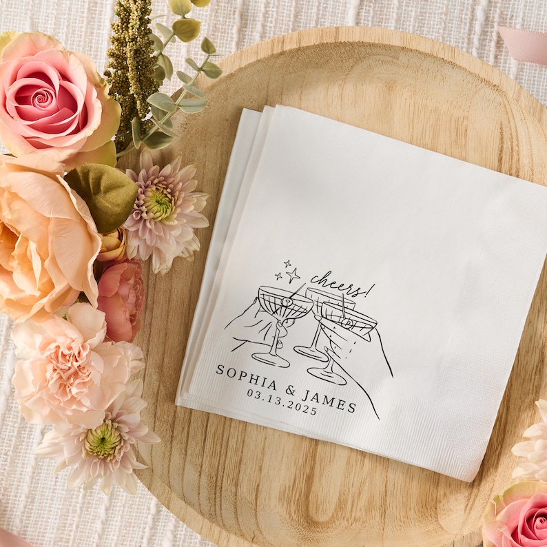 K&ouml;nnte beinhalten: Eine wei&szlig;e Papierserviette mit einer schwarzen Strichzeichnung von zwei H&auml;nden, die Martinigl&auml;ser zum Ansto&szlig;en halten. Der Text lautet cheers! gefolgt von den Namen SOPHIA & JAMES und dem Datum 13.03.2025 in einer schwarzen Serifenschrift.