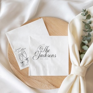 K&ouml;nnte beinhalten: Zwei quadratische wei&szlig;e Papierservietten mit schwarzem Aufdruck. Eine Serviette zeigt den Text "Cheers to The Jacksons EST. 2026" in Schreibschrift. Die andere Serviette zeigt zwei Sektgl&auml;ser mit Schleifen und den Text "Denise + Matthew".