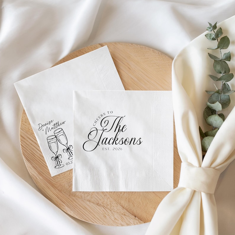 K&ouml;nnte beinhalten: Zwei quadratische wei&szlig;e Papierservietten mit schwarzem Aufdruck. Eine Serviette zeigt den Text "Cheers to The Jacksons EST. 2026" in Schreibschrift. Die andere Serviette zeigt zwei Sektgl&auml;ser mit Schleifen und den Text "Denise + Matthew".