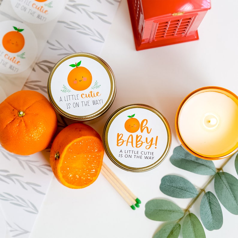 Op de afbeelding: Twee gouden kaarsenpotten met oranje geurkaarsen. De etiketten op de potten hebben een schattige sinaasappelillustratie en de tekst "A little cutie is on the way!" en "Oh Baby! A little cutie is on the way".