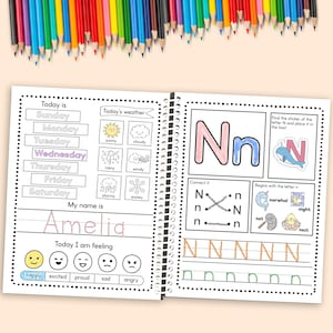 May include: A spiral-bound activity book open to two pages, featuring colorful illustrations and text. The left page includes sections for writing the day, weather, name, and feelings. The right page focuses on the letter "N" with tracing exercises and related images. Colored pencils are scattered above.