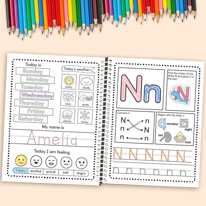 May include: A spiral-bound activity book open to two pages, featuring colorful illustrations and text. The left page includes sections for writing the day, weather, name, and feelings. The right page focuses on the letter "N" with tracing exercises and related images. Colored pencils are scattered above.