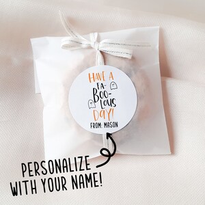 Op de afbeelding: Een witte ronde sticker met oranje tekst die "Have a Fa-Boo-Lous Day!" luidt, met twee geestillustraties. De tekst "From: Mason" staat onder de hoofdtekst. De sticker is bevestigd aan een witte papieren zak met een zilveren lint.